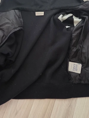 Svart pufferjacka från Moncler - Säljer en svart pufferjacka från Moncler med stor vit M-detalj på ärmen och Moncler-logga. Jackan har huva och quiltade partier samt stickade muddar. Materialet är en mix av vadderad nylon och stickad ull, perfekt för kalla vinterdagar.