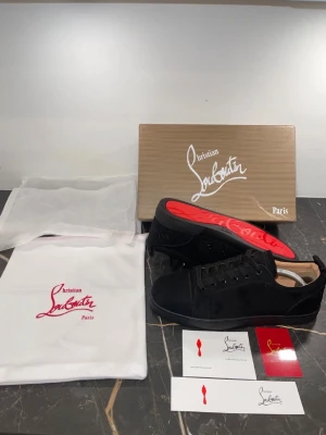Svarta sneakers från Christian Louboutin - Stilrena svarta sneakers från Christian Louboutin i mocka med klassisk röd sula. Skorna har svarta snören, diskreta detaljer och en låg profil. Perfekta för dig som vill ha en lyxig och ikonisk look med en touch av Paris.