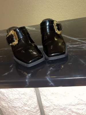 Svarta loafers Versace Jeans Couture - Säljer ett par svarta loafers från Versace Jeans Couture i lackat skinn. Skorna har fyrkantig tå och en bred rem med en stor, dekorativ guldfärgad spänne med märkets logga. Riktigt statement-plagg för dig som gillar lyxiga detaljer.