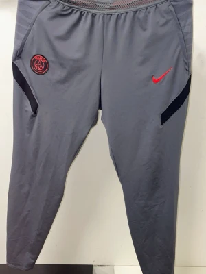 Gråa PSG träningsbyxor Nike - Snygga gråa träningsbyxor från Nike med PSG-logga och röd Nike Swoosh på framsidan. Byxorna har smal passform, elastisk midja och svarta detaljer vid fickorna. Perfekta för fotbollsträning eller gymmet.