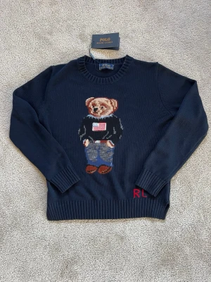 Mörkblå stickad tröja Polo Bear - Mörkblå stickad tröja från Polo Ralph Lauren med ikoniska Polo Bear-motivet framtill. Björnen har på sig jeans och en svart tröja med amerikanska flaggan. Tröjan har ribbade muddar och rund hals. Perfekt för dig som gillar klassisk preppy stil.