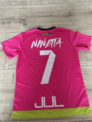 Rosa sporttröja JUL x Ninjetta - Snygg rosa sporttröja från JUL x Ninjetta med tryckta detaljer i vitt, svart och limegrönt. Nummer 7 på ryggen och coola grafiska mönster framtill. Tillverkad i lätt och snabbtorkande material, perfekt för träning eller match.