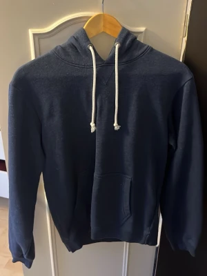 Mörkblå hoodie från Pier One - Mörkblå hoodie från Pier One med klassisk känguruficka och vita dragsnören i huvan. Hoodien har en enkel och stilren design, perfekt för en avslappnad look. Tillverkad i mjukt material som känns skönt mot huden. Lite trasig vid hakan men går att sy så de inte syns