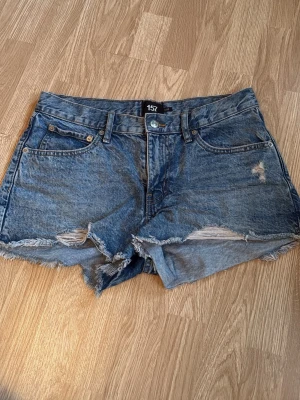 Blå jeansshorts från 157 med slitningar - Säljer ett par blå jeansshorts från 157 med råa kanter och slitna detaljer på framsidan. I storlek M.