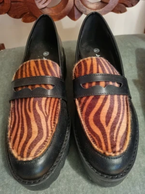 Zebramönstrade loafers Monki - Unika loafers med svart konstskinn och zebramönstrad ovandel i bruna och svarta toner. Snygg detalj med bred rem över foten och chunky sula som ger extra edge. Perfekta för dig som vill sticka ut med din stil. Aldrig använda, så i nyskick. 