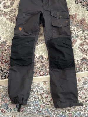 Svarta cargobyxor Fjällräven dam - Snygga svarta cargobyxor från Fjällräven i dammodell, storlek 38. Byxorna har flera praktiska fickor, förstärkta knän i svart tyg och justerbara benslut. Tillverkade i slitstarkt material, perfekta för äventyr och outdoor.