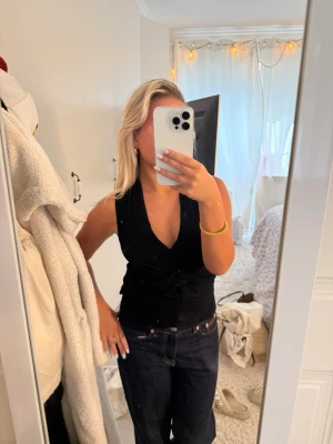 Zara Svart ärmlös omlottopp - Snygg svart omlottopp med djup v-ringning och knyt i midjan. Toppen är ärmlös och har en figurnära passform som framhäver siluetten. Perfekt att styla med jeans eller kjol för en trendig look.