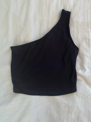 Svart oneshoulder topp - Snygg svart oneshoulder topp i stretchigt material. Toppen är croppad och har en asymmetrisk axelbandslös design som ger en trendig look. Perfekt att matcha med höga jeans eller kjol för en cool stil.