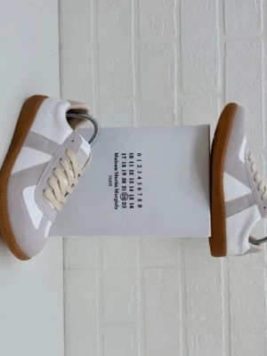 Maison Margiela vita sneakers Storlek 43 - Maison Margiela sneakers i vitt och ljusgrått med beige mockadetaljer och klassiska gum-yttersulor. Låga sneakers med retrodesign, tillverkade i läder och mocka. Snygga och stilrena med beige snörning och ikonisk Margiela-box. Storlek 43 och de är i helt nyskick. Pris kan diskuteras 🤙