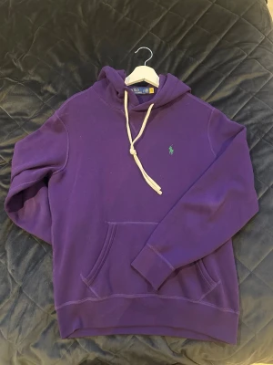 Lila hoodie från Polo Ralph Lauren - Snygg lila hoodie från Polo Ralph Lauren med klassisk huva och vita dragsnören. Har en broderad grön logga på bröstet och en stor känguruficka framtill. Perfekt för en avslappnad och trendig stil. Tillverkad i mjukt bomullsmaterial.