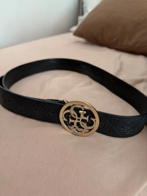 Svart skärp med guldspänne från Guess - Snyggt svart skärp från Guess med präglat mönster längs hela bandet och ett stort, runt guldspänne med Guess-logga. Perfekt accessoar för att lyfta vilken outfit som helst. Skärpet har en klassisk och trendig look.
