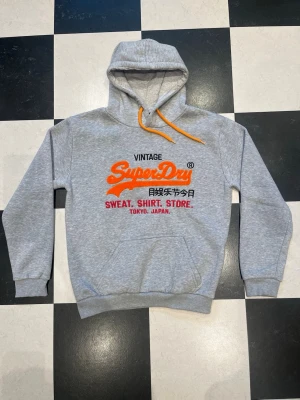 Grå Superdry hoodie med tryck - Snygg grå hoodie från Superdry med stor logga och text i orange, svart och rött på bröstet. Klassisk känguruficka framtill, ribbade muddar och huva med orange dragsko. Tillverkad i mjuk bomullsblandning, perfekt för en avslappnad streetwear-look. Tröjan är smått nopprig över hela men inte så synligt när man när den