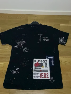 Dsquared2 tröja svart. - Svart DSQ2-inspirerad T-shirt med unik design – endast 1 kvar i lager.  Den här tröjan sticker verkligen ut med sina färgstänk (paint splatter) och grafiska tryck framtill. Perfekt för en streetwear look eller som statement piece i en annars enkel outfit.  ✔️ Färg: Svart med vita/färgade detaljer ✔️ Stil: Oversized / relaxed fit ✔️ Design: Grafiskt tryck + DSQ2 patch framtill ✔️ Skick: [skriv här: ny / mycket bra skick] ✔️ Storlek: [lägg till storlek här]  💥 Endast 1 kvar – säljs inte igen 💥 Passar s