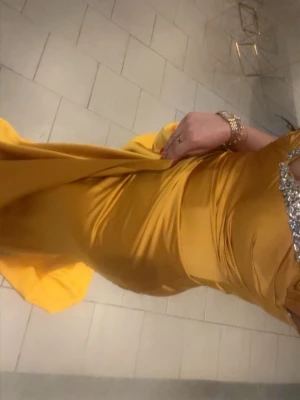 Gul långklänning med slits och glitter - Snygg gul långklänning med glittriga detaljer vid bysten och en djup slits på sidan. Klänningen har offshoulder-ärmar och är gjord i ett glansigt satinliknande material som ger en lyxig känsla. Perfekt för dig som vill sticka ut och glänsa.