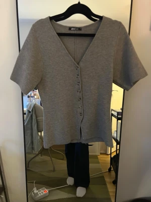 Grå kortärmad kofta från Gina Tricot - Grå kofta från Gina Tricot med v-ringning och knappar framtill. Kortärmad och i mjukt stickat material. Storlek M och säljer då den inte kommer till användning. Skriv för frågor!💕