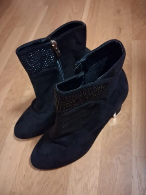 Svarta stövletter med gulddetalj - Snygga svarta stövletter med chunky klack och guldfärgad detalj på hälen. Ovandelen har dekorativa svarta strass och dragkedja på sidan. Materialet ser ut att vara mocka eller liknande. Perfekt för dig som vill ha en cool och trendig look.