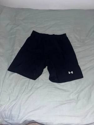 Svarta träningsshorts från Under Armour - Säljer ett par svarta träningsshorts från Under Armour med elastisk midja och snörning. Shortsen har en loose fit och är tillverkade i ett lätt och snabbtorkande syntetmaterial. Perfekta för gymmet eller löprundan.
