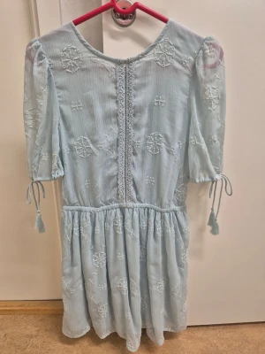 Ljusblå broderad klänning med snörning 34 - Supersöt ljusblå klänning med broderade blommor, puffärmar med knytband och tofsar samt vacker snörning i ryggen. Klänningen har en lös passform och fina spetsdetaljer framtill. Perfekt för sommarens alla tillfällen!