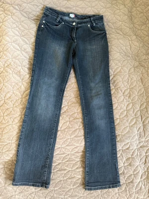 Blå bootcut jeans från Outfit Fashion - Bootcut jeans i klassisk blå denim från Outfit Fashion. Jeansen har fem fickor, silverfärgade knappar och subtila slitningar. Materialet är bomullsdenim och passformen är rak med lätt utsvängda ben. Perfekt för dig som gillar en tidlös och avslappnad look.