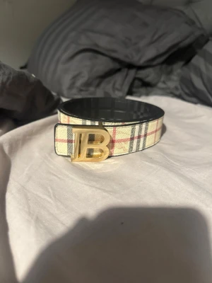Rutig Burberry-bälte med gulddetalj - Snyggt bälte från Burberry med klassiskt rutigt mönster i beige, svart och rött. Bältet har ett stort guldfärgat 'TB'-spänne som ger en exklusiv känsla. Perfekt accessoar för att lyfta vilken outfit som helst.