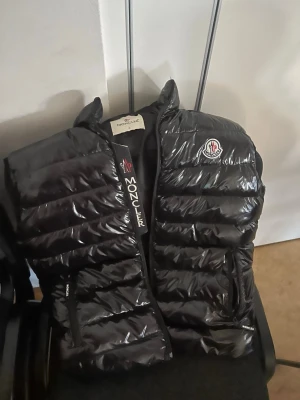 Svart dunväst från Moncler - Snygg svart dunväst från Moncler med glansigt material och klassisk logotyp på bröstet. Västen har en rak passform, dragkedja framtill och diskreta Moncler-detaljer. Perfekt för lager-på-lager och håller dig varm under kyliga dagar.