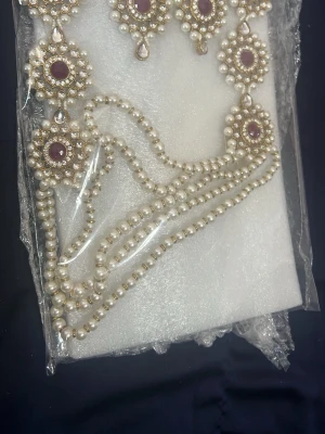 Elegant halsband med pärlor och stenar - Säljer ett elegant halsband med vita pärlor och dekorativa medaljonger med lila stenar och gnistrande kristaller. Halsbandet har flera pärlsträngar och detaljerade blomliknande former. Perfekt för dig som gillar unika accessoarer.