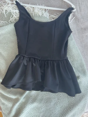 Svart peplumtop med volang - Snygg svart peplumtop med breda axelband och volang nedtill. Toppen har en figurnära passform upptill och är tillverkad i ett slätt syntetmaterial som ger en elegant look. Perfekt att matcha med jeans eller kjol för en trendig stil.