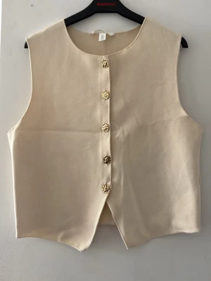 Beige topp med guldfärgade knappar - Stilren beige topp med rund halsringning och fem unika guldfärgade knappar framtill. Toppen har en rak och enkel form med slits nedtill. Materialet är mjukt och slätt, perfekt för att skapa en clean look. Denna topp har aldrig använts innan, den är otroligt stilig och perfekt för dig som tycker om att vara snygg i din vardag eller vid större event