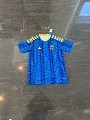 Blå Sverige tröja från Adidas - Säljer en blå matchtröja från Adidas med Sveriges landslagsemblem på bröstet. Tröjan har gula detaljer på krage och axlar, samt ett unikt mönster i olika blå nyanser. Materialet är lätt och andas, perfekt för sport och träning.