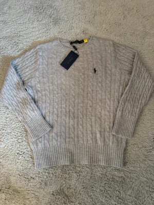 Polo Ralph Lauren tröja  - Snygg ljusgrå kabelstickad tröja med rund halsringning och klassisk broderad logga på bröstet👌😍. Tröjan har långa ärmar och ribbade muddar vid ärmslut och nederkant. Perfekt för lager på lager och stilren look. Själv är jag 176 och den är precis lite för kort och tajt. Måtten finns med på bilder, du får med lappen fast den är avklippt.