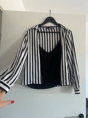 Randig svartvit blus med lång ärm - Snygg svartvit blus med vertikala ränder och långa ärmar. Blusen har en klassisk krage och knäppning framtill. Passar perfekt till både jeans och kjol för en trendig look. Materialet känns lätt och luftigt.