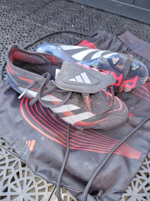 Adidas Predator fotbollsskor svart/röd - Säljer ett par Adidas Predator fotbollsskor i svart med röda och vita detaljer. Skorna har snörning, klassisk Predator-tunga och mönstrad sula för bra grepp på planen. Kommer med tillhörande svart och röd skopåse. Perfekta för dig som vill dominera på fotbollsplanen. Använda 5 ggr. FG dubb.