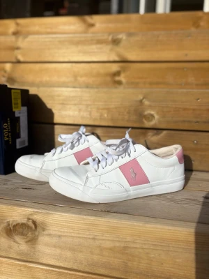 Vita och rosa Polo Ralph Lauren sneakers - Snygga vita sneakers från Polo Ralph Lauren med rosa detaljer på sidan och hälen. Klassisk låg modell med vita skosnören och Polo-logga broderad på hälen. Snygga och fräscha skor😍 skosnörena är lite smutsiga men går att tvätta. Se bild för skorna fram, lite skav men inget som märks i helhet! Original kartong får man med. Hör av er vid intresse, pris eller fler bilder!🩷nypris 1000kr
