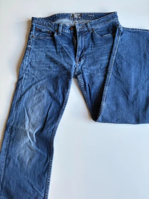 Blå jeans från Dressmann 33/30 - Säljer ett par klassiska blå jeans från Dressmann i storlek 33/30. Jeansen har fem fickor, normal passform och är gjorda i slitstarkt denimtyg. Perfekta för en avslappnad och stilren look. Snygga detaljer med kontrastsömmar och knappgylf.