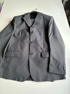 Grå randig kavaj + byxor Made in Italy - Snygg grå kavaj med tunna vita ränder, klassisk passform och tre knappar framtill. Kavajen har två fickor med lock och en bröstficka. Perfekt för dig som vill ha en stilren och tidlös look. Strl  M/L helt kostym set med påse byxor och kavaj