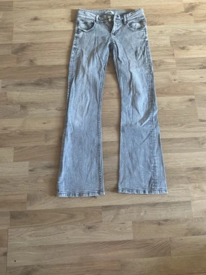 Grå bootcut jeans från Gina Tricot - Säljer ett par grå bootcut jeans från Gina Tricot med dubbla silverknappar och klassisk femficksmodell. Jeansen har en lätt utsvängd passform och är tillverkade i denim med snygg tvättad look. Perfekta för dig som gillar retrostil och vill ha ett par bekväma jeans.