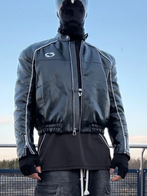 Svart skinnjacka från Oxbow - Cool svart skinnjacka från Oxbow med vita detaljer och logga på bröstet. Jackan har dragkedja framtill, elastiska muddar vid ärmslut och nederkant samt två fickor. Perfekt för dig som vill ha en edgy och sportig look.