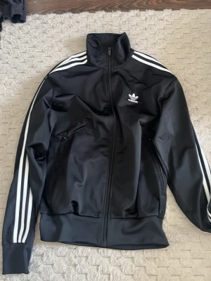Svart Adidas track jacket - Säljer en svart Adidas track jacket med klassiska vita ränder längs ärmarna och Adidas logga på bröstet. Jackan har hög krage, dragkedja framtill och två fickor med dragkedja. Perfekt för sport eller chill. Materialet är polyester.