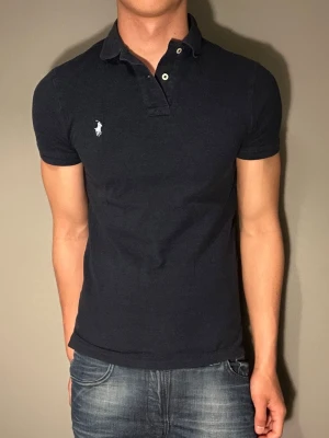 Mörkblå piké från Polo Ralph Lauren - Klassisk svart polotopp från Polo Ralph Lauren med vit broderad logga på bröstet. T-shirten har korta ärmar, krage och knappar framtill. Materialet är bomull och passformen är slim. Perfekt för dig som gillar stilrena och tidlösa plagg.