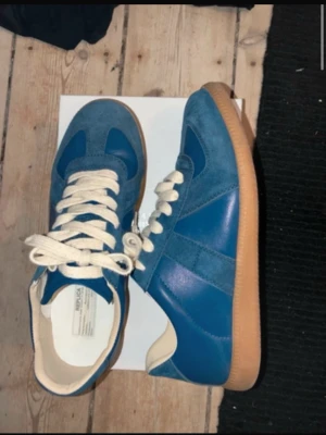 Blå sneakers från Maison Margiela Storlek 44 - Säljer ett par blå sneakers från Maison Margiela med beige sula och vita skosnören. Skorna har en mix av mocka och läder på ovandelen och klassisk låg siluett. Perfekta för dig som gillar stilrena och trendiga sneakers. Storlek 44 och är i nyskick och aldrig använda ,pris kan diskuteras, hör av dig 🤙. Ni vet varför priset är så lågt 👍👍