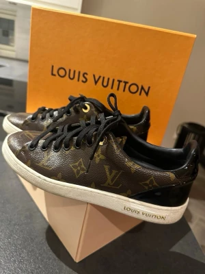 Louis Vuitton bruna sneakers - Snygga bruna sneakers från Louis Vuitton med klassiskt monogrammönster i guld och svart. Skorna har svarta skosnören, vit sula med Louis Vuitton-logga och är tillverkade i läder med exklusiv känsla. Perfekt för dig som vill sticka ut med lyxiga detaljer.
