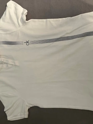 Vit t-shirt från Calvin Klein - Vit piké tröja från Calvin Klein med korta ärmar och diskret CK-logga samt svarta linjer på framsidan. Klassisk passform och knappar vid halsen. Perfekt för dig som gillar stilrena plagg.
