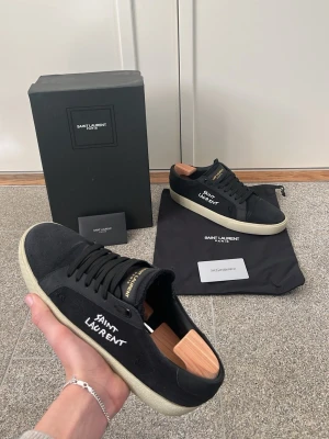 Saint Laurent svart låga sneakers - Svarta sneakers från Saint Laurent med vit text på sidan och guldfärgade detaljer på plösen. Skorna har en klassisk låg profil, vit sula och är tillverkade i canvas med läderdetaljer. Perfekt för dig som vill ha en stilren och exklusiv look.