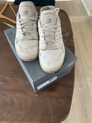 New Balance 550 vita sneakers - Säljer ett par vita New Balance 550 sneakers med grå detaljer. Klassisk låg modell med skön passform och robust sula. Materialet är en mix av läder och textil, med tydliga New Balance-loggor på sidan och tungan. Perfekta för dig som gillar stilrena och sportiga skor.
