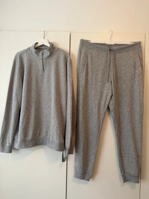 Grått loungewear-set med dragkedja - Mysigt grått loungewear-set med långärmad tröja med halv dragkedja och hög krage samt matchande byxor med dragsko i midjan och muddar vid benslut. Tillverkat i mjukt, stretchigt material som passar perfekt för chill dagar hemma.