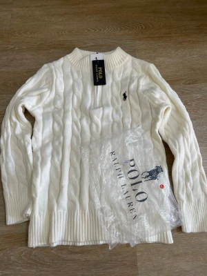 Vit kabelstickad tröja Polo Ralph Lauren - Vit kabelstickad tröja från Polo Ralph Lauren med klassisk svart logobrodyr på bröstet. Tröjan har ribbstickad krage, ärmslut och nederkant. Perfekt för dig som gillar stilrena och tidlösa plagg.