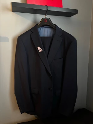 Två kavajer från dressman - Stilren mörkblå kavaj från Dressmann med klassisk passform och snygga detaljer. Kavajen har två knappar framtill, flera innerfickor med röda kantband och ljusblå foder. Perfekt för dig som vill ha en enkel och elegant look. Ena är storlek 48 och den andra storlek 56. Skriv till mig vilken du är intresserad i. Byxor: 32/32 Kavaj 1: 56 kavaj 2: 48