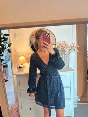 Marinblå omlottklänning med glitter - Kolla min vinted istället!! Snygg marinblå kort omlottklänning med glittriga detaljer och knytband i midjan. Klänningen har långärmade ärmar och v-ringad hals, perfekt för dig som vill ha en trendig look med lite extra shine.