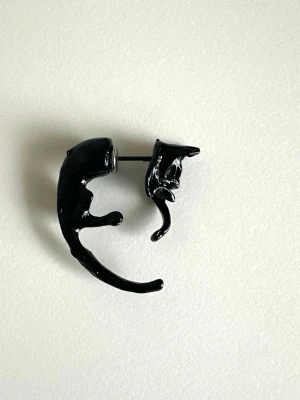 Svart kattörhänge i metall - Unikt svart örhänge format som en katt som slingrar sig runt örsnibben. Örhänget är gjort i metall och har en cool, lekfull design som verkligen sticker ut. Perfekt för dig som gillar djurmotiv och vill ha något annorlunda i smyckeskrinet.