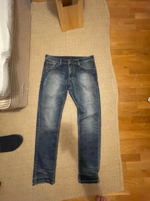 Nudie jeans me slitningar - Säljer ett par blå nudie jeans med snygga slitningar och ljusa detaljer på benen. Klassisk femficksdesign och knappstängning. Jeansen har en modern passform och är tillverkade i denim med stretch för extra komfort. Modellen är oklar men de straight/ slim fit strl W32 L33